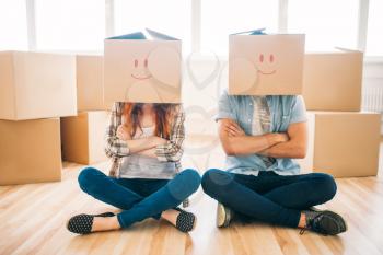Playful couple with cardboard boxes on their heads sitting on the floor, moving to new home, housewarming. Man and woman have a fun