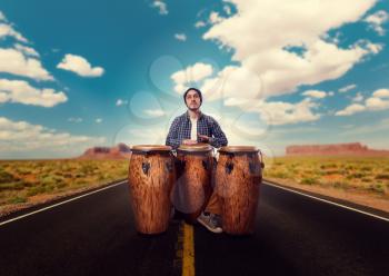Young male drummer with wooden bongo drums plays on desert road, musician in motion. Djembe, musical percussion instrument, ethnic music