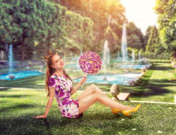 Young pretty girl holding a huge colorful lollypop. Amasing young woman sits on green grass holding big candy in her hands. Stylish girl in summer dress, park with fountains on background.