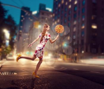 Beautiful young woman run with huge lollipop in her hand. Stylish girl with blonde curly hair. Stylish girl in colorful summer dress, night city on background.