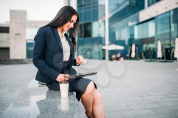 Businesswoman in suit works on laptop outdoor. Modern building, financial center, cityscape. Female businessperson working