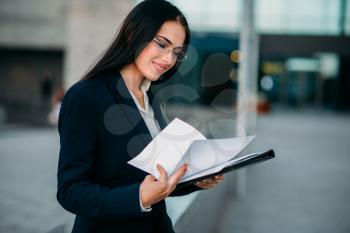 Businesswoman in suit works with notebook outdoor, business center on background. Modern financial building, cityscape. Successful female businessperson