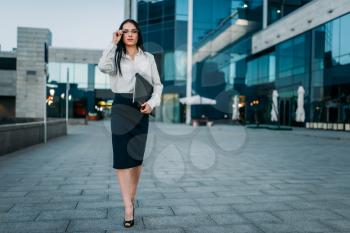 Portrait of young business woman in glasses and white blouse with notebook in hand, skyscaper on background. Modern building, financial center, cityscape. Successful female businessperson