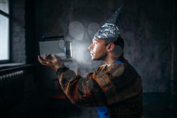 Paranoid man in tinfoil cap watch TV, mind protection from telepathy, paranoia concept. UFO phobia
