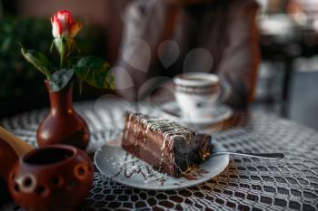 Delicious chocolate cake and a cup of coffee on table in cafe. Table decorated with white knitted tablecloth and vase with rose. Woman sits at table in cafe, selective focus.
