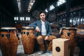 Young male drummer pose against african wooden drum, factory shop on background. Djembe, musical percussion instrument, bongo beats