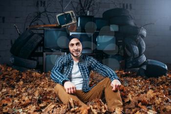 Stalker, male traveler sitting on the ground, the mountain of old TVs on background, remains of civilization. Danger zone, post apocalypse horror