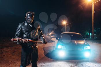 Serial maniac in leather coat and hockey mask, bloody baseball bat wrapped in metal chain in hands, against black car with light at the night. Horror, bloody murderer, murder weapon