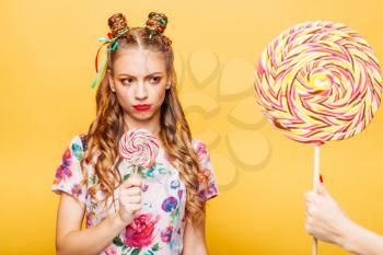 Beautiful young woman with mistrustful look holds in hand big lollypop. Somebody gives one more candy to girl, studio portrait. Stylish girl in summer colorful dress, yellow wall on background.