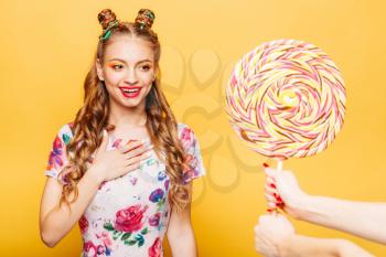 Beautiful young woman surprised somebody gives her huge lollypop. Bright girl with blonde curly hair. Stylish girl in summer colorful dress, yellow wall on background.