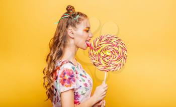 Beautiful young woman with playful look eating huge candy and smiling. Stylish girl with blonde curly hair. Portrait of attractive lady with big lollypop, yellow wall on background.
