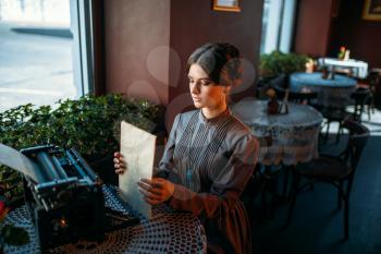 Happy young woman sits by the table in cafe and looks thoughtfully out the window. Portrait of retro style woman in vintage cafe with typewriter. Vintage style cafe, vase with rose on a table.