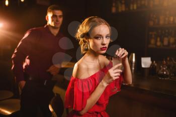 Woman in red dress with cocktail in hand, man behind bar counter, flirting. Date in nightclub, attractive love couple in pub