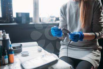 Female tattooer in blue sterile gloves prepares tattoo machine, professional work tools on background. Tattooing in salon