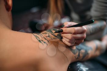 Female tattooist drawing tattoo on male shoulder. Professional tattooing in salon