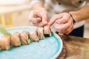 Male person cooking sushi rolls with salmon on wooden table, japanese food preparation process.