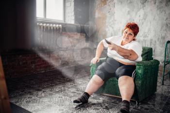 Overweight woman sits in a chair and watches TV. Unhealthy lifestyle, obesity