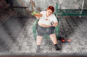 Overweight woman with sandwich in hands sits in a chair and watches TV, bulimic, fatty. Unhealthy lifestyle, obesity