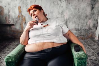 Overweight woman sits in a chair and eats sweet cake, laziness and obesity, bulimic. Unhealthy food eating, fatty female