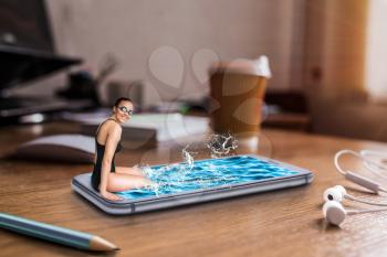 Female swimmer sits on the edge of large phone screen with water, table with big coffee cup on background. Mobile and communication technology addiction, social addicted people. Scaling effect