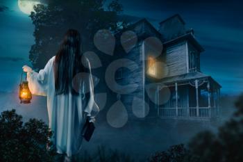 Female person in white shirt holds kerosene lamp and spellbook in hand against abondoned house, moonlit night, back view. Dark magic, occultism and exorcism