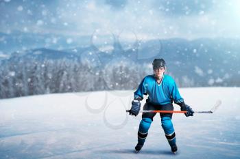One hockey player with stick on open ice, game concept, snowy forest on background. Male person in helmet, gloves and in protective uniform outdoors, winter sports game