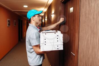 Pizza delivery boy knocking on the door of the customer, delivering service. Courier from pizzeria holds cardboard packages indoors