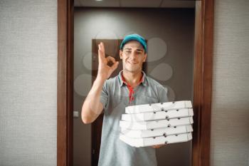 Cheerful pizza delivery boy shows Ok symbol, delivering service. Courier from pizzeria holds cardboard packages indoors
