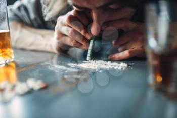 Male junkie sniffing a line of cocaine, grugs and bottle of alcohol on the table on background. Addiction concept, addicted people