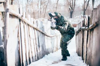 Male paintball player with marker gun in hands attack, front view, winter battle. Extreme sport game, soldier fights in protection mask and uniform