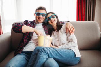 Smiling couple in 3D glasses sitting on couch and watch tv with popcorn at home, man with remote control in hand, window on background