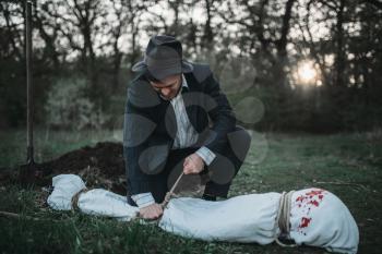 Maniac ties a rope on the victim's body wrapped in a canvas, grave on background, serial murderer concept, crime horror