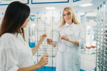 Female optician shows glasses to buyer in optics store. Selection of eyeglasses with professional optometrist