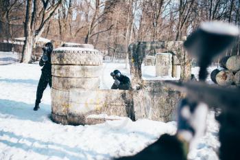 Paintball player hands with marker gun shooting at the enemy, winter forest battle. Extreme sport game