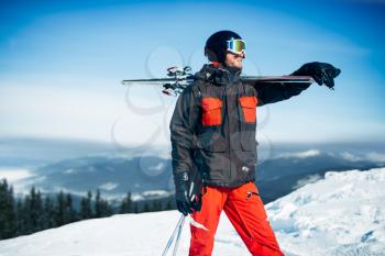 Skier poses with skis and poles in hands, blue sky and snowy mountains on background. Winter active sport, extreme lifestyle. Downhill skiing