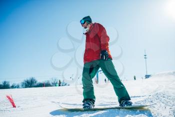 Snowboarder in glasses poses with board in hands, blue sky and snowy mountains on background