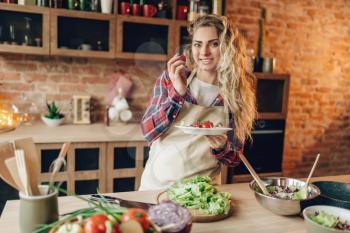Smiling young housewife in apron prepares salad, kitchen interior on background. Female cook making healthy vegetarian food, vegetables preparation