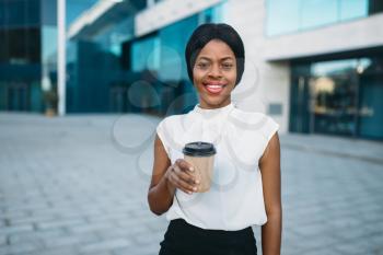 Smiling business woman with cardboard coffee cup outdoors, office building on background. Black businesswoman in skirt and white blouse