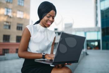 Young business woman uses laptop against office building. Smiling black businesswoman in skirt and white blouse works outdoors