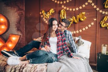 Happy young couple hugs on the bed in bedroom with lighting decoration. Man and woman in clothes embrace on couch