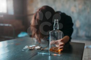 Female junkie with bottle of alcohol sitting at the table with drugs and syringe, grunge room interior on background. Drug addiction concept, addicted people