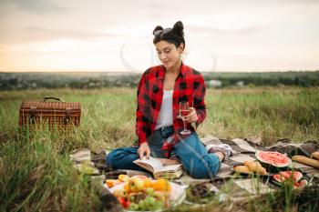 Cute woman with glass of wine reads book, picnic on meadow. Romantic junket, happy holiday