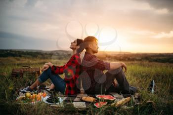 Love couple sitting with their backs to each other on sunset, picnic in the field. Romantic junket on sunset, man and woman on outdoor dinner,  happy family weekend
