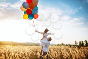 Happy love couple with balloons hugs in wheat field. Pretty wife and husband walking on summer meadow