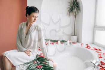 Elegant woman in white bathrobe sitting on the edge of the bath decorated with rose petals. Luxury bathroom interior