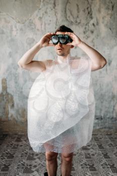 Naked freak man wrapped in packaging film wearing swimming goggles, grunge room interior. Crazy male person in abandoned house