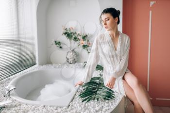 Beautiful woman in white bathrobe sitting on the edge of the bath with foam. Bathroom interior with window on background