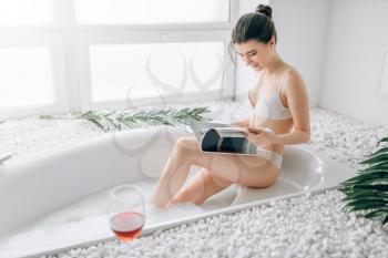 Attractive woman in white underwear reading magazine and relax in bath, glass of red wine standing on the edge. Luxury bathroom interior, stone decor