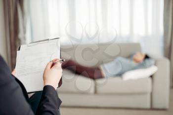 Female patient lies on sofa at psychotherapist reception. Female doctor writes notes in notepad, professional psychology support