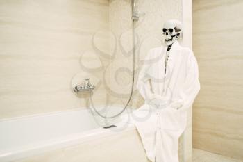 Human skeleton in white bathrobe sitting on the edge of the bath in bathroom, black humor